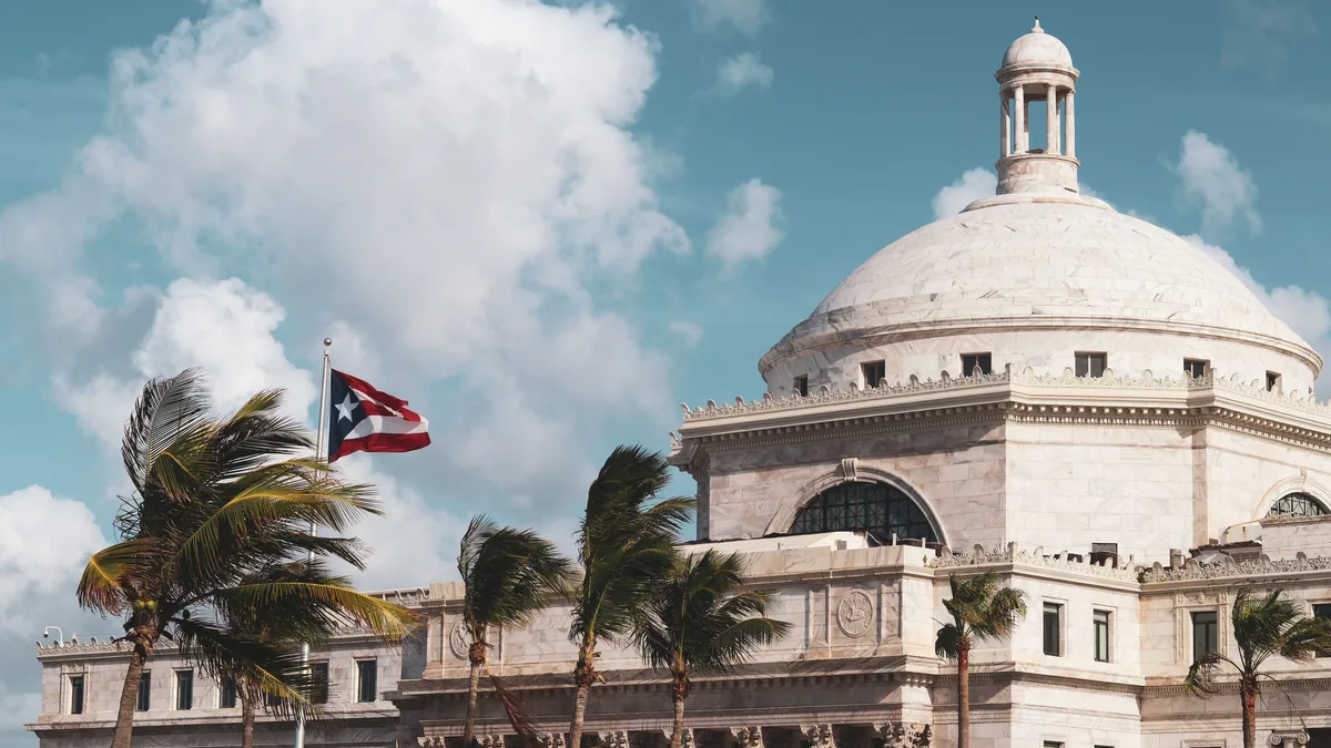 The Puerto Rico Capitol building with a Puerto Rican flag flying in front of it, surrounded by palm trees and a blue sky with clouds.