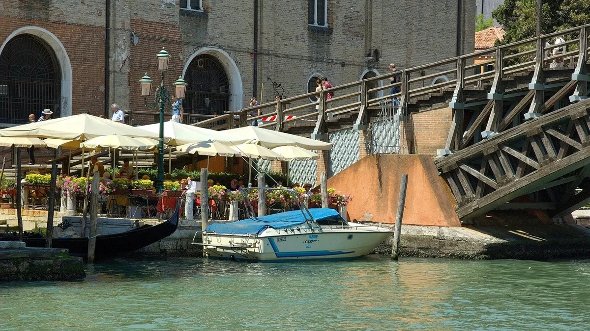 A boat docked at a canal in Venice, Italy, with a bridge and buildings in the background.