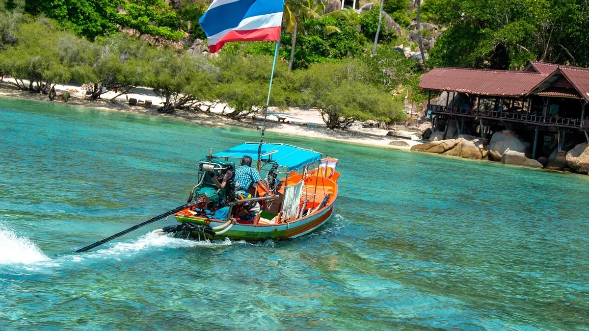 A long-tail boat with a Thai flag flying from its bow, traveling through turquoise water near a beach with a building in the background.