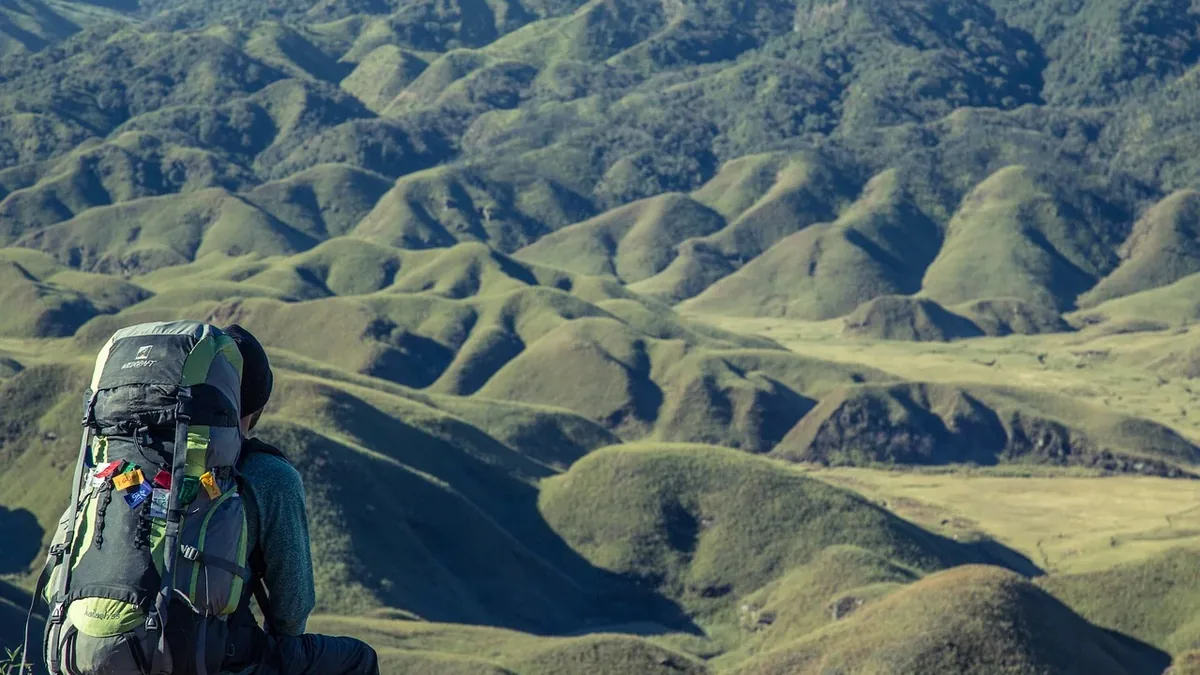 A person wearing a large backpack, sitting on a hillside with rolling hills and mountains in the background.